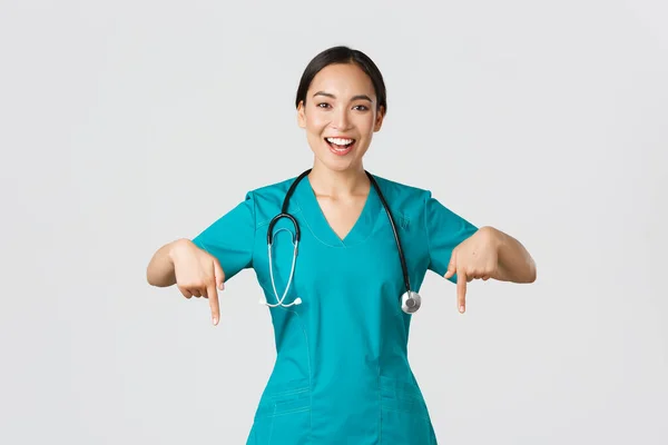 Covid-19, profissionais de saúde, conceito pandémico. Feliz sorrindo bela enfermeira asiática, médico feminino em esfrega apontando para baixo, convidando para check-up, mostrando anúncio do hospital, fundo branco — Fotografia de Stock