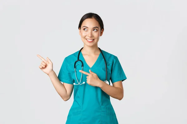 Covid-19, Gesundheitspersonal, Pandemiekonzept. Fasziniert schöne asiatische Praktikantin, Krankenschwester in Peelings suchen und zeigen oben links Ecke mit neugierig verträumtes Lächeln, weißer Hintergrund — Stockfoto
