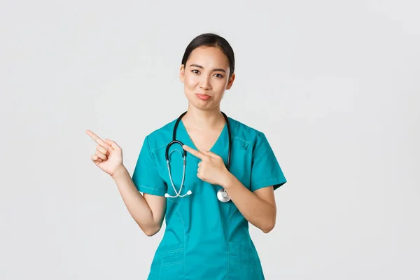 Covid-19, egészségügyi dolgozók, világjárvány. Szkeptikus és kétes ázsiai női ápolónő köpenyben rámutatva a bal felső sarokban, és grimaszolva szórakozás nélkül, nem hasonló termék, hogy tétova — Stock Fotó