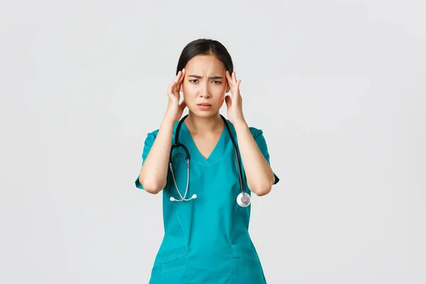 Covid-19, egészségügyi dolgozók, világjárvány. Túlhajszolt és kimerült ázsiai női orvos, hányinger, köpeny viselése, fejérintés, fejfájás vagy magas láz panasza — Stock Fotó