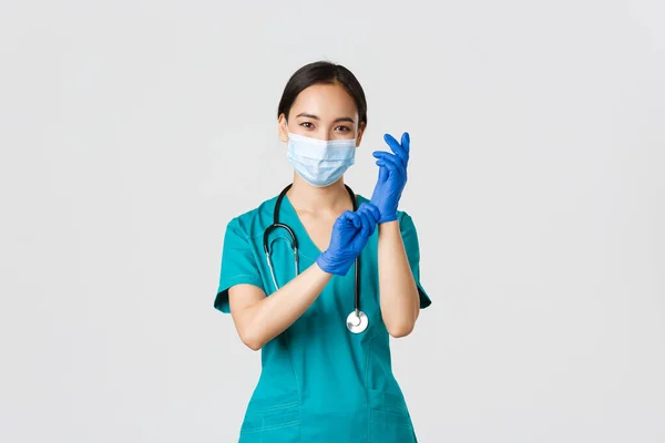 Covid-19, koronavirüs hastalığı, sağlık çalışanları konsepti. Profesyonel ve kendinden emin gülümseyen Asyalı doktor, hemşire hasta muayenesi için lastik eldiven ve tıbbi maske takıyor. — Stok fotoğraf