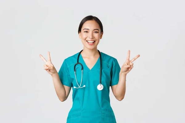 Covid-19, sjukvårdspersonal, pandemikoncept. Glad asiatisk kvinnlig läkare, sjuksköterska i buskar visar fred kawaii gest och leende, vistas positiv under arbetet på sjukhus, vit bakgrund — Stockfoto