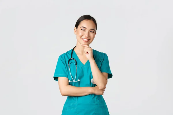 Covid-19, healthcare workers, pandemic concept. Smiling pleased attractive asian female doctor in scrubs, looking upper left corner and thinking, have idea, standing thoughtful white background — Stock Photo, Image