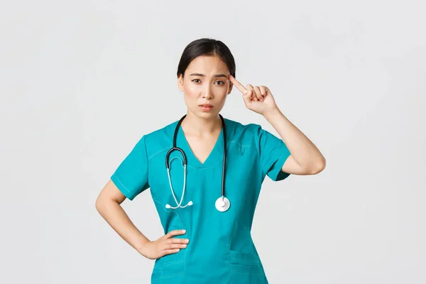 Covid-19, healthcare workers, pandemic concept. Annoyed skeptical asian female nurse or doctor scolding someone acting crazy or stupid, roll finger over temple with judgemental face — Stock Photo, Image