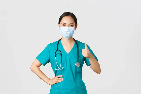 Covid-19, koronavírus betegség, egészségügyi dolgozók koncepciója. Magabiztos mosolygós ázsiai orvos, női nővér vagy orvos orvosi maszk és köpeny, mutassa hüvelykujj-up biztosítja a minőséget, guanratee szolgáltatás — Stock Fotó