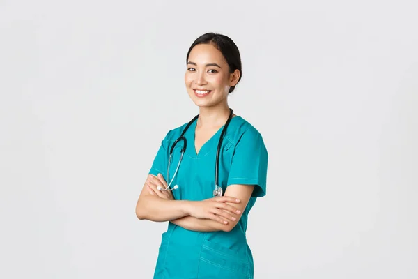 Covid-19, sağlık çalışanları, salgın konsepti. Profesyonel özgüvenli, gülümseyen Asyalı kadın doktor, muayene için önlüklü hemşire, çapraz kollar ve kameraya bakın. — Stok fotoğraf