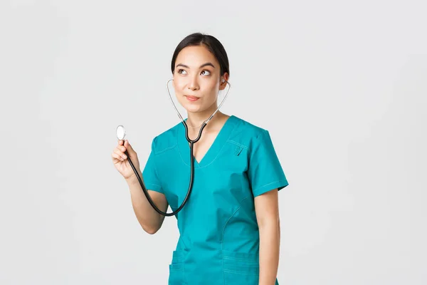 Covid-19, healthcare workers and preventing virus concept. Young focused asian female doctor, physician in scrubs, examine patient lungs, using stethoscope, standing thoughtful white background — Stock Photo, Image