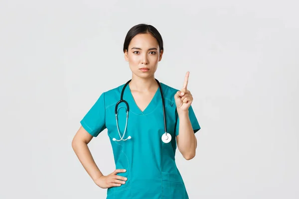Covid-19, zdravotníci, pandemický koncept. Vážně vypadající profesionální asijská lékařka v drhnutí, lékař třesoucí ukazováčkem v prohibici, varovné gesto, něco zakázat — Stock fotografie