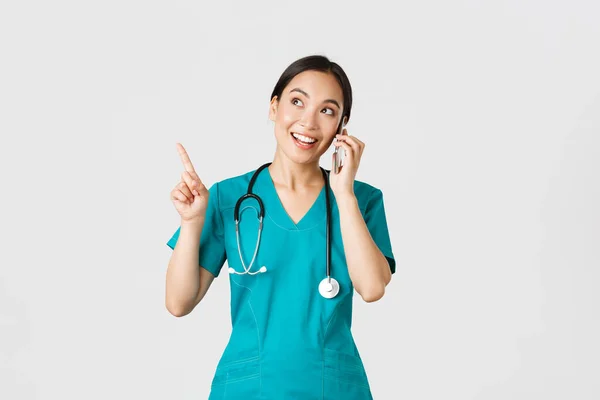 Covid-19, profissionais de saúde e prevenção do conceito de vírus. Retrato de jovem muito asiático médico feminino, enfermeira em esfoliantes fazendo ordem on-line, falando por telefone e apontando canto superior esquerdo, sorrindo — Fotografia de Stock