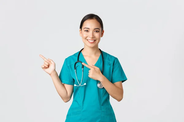 Covid-19, profissionais de saúde, conceito pandémico. Sorrindo muito asiático médico, enfermeira em esfrega e estetoscópio, apontando os dedos canto superior esquerdo, mostrando banner clínica on-line, fundo branco — Fotografia de Stock