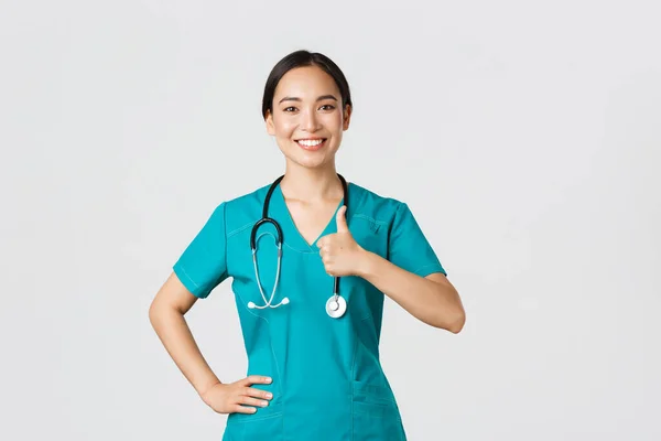 Covid-19, healthcare workers, pandemic concept. Smiling upbeat, confident female asian nurse in scrubs have all under control, show thumbs-up in approval, guarantee quality of service in clinic — Stock Photo, Image