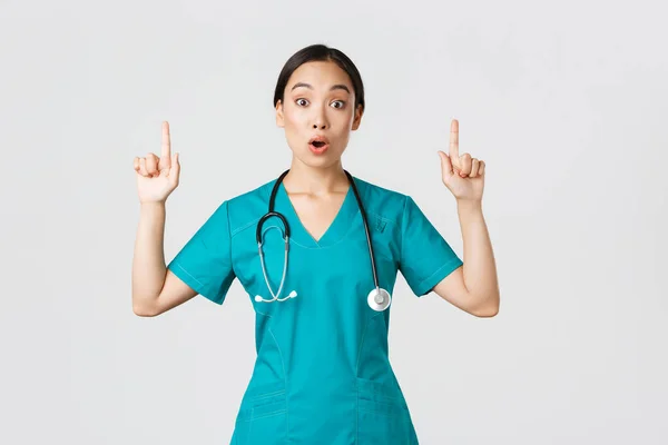 Covid-19, sağlık çalışanları, salgın konsepti. Şaşırmış ve heyecanlanmış Asyalı kadın hemşire, önlüklü kadın doktor ilginç bir promosyon soruyor, parmakla işaret ediyor, klinik reklamını gösteriyor. — Stok fotoğraf