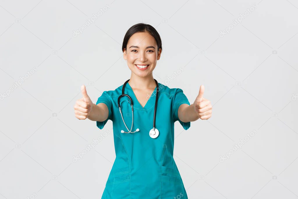 Covid-19, healthcare workers, pandemic concept. Smiling pretty asian female doctor, nurse in scrubs ensure everything good, showing thumbs-up in approval, agree or like something, white background