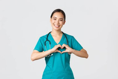 Covid-19, sağlık çalışanları, salgın konsepti. Sevecen Asyalı doktor, önlüklü kadın hemşire kalp hareketi ve gülümseme gösteriyor, beyaz geçmişi olan hastalarla ilgileniyor.
