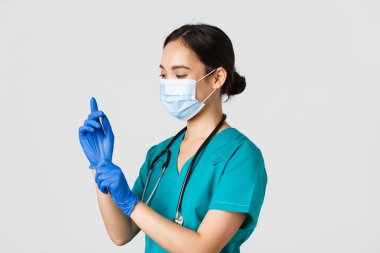 Covid-19, koronavirüs hastalığı, sağlık çalışanları konsepti. Profesyonel gülümseyen Asyalı hemşire, ameliyat önlüğü ve tıbbi maske giyen doktor muayene için lastik eldiven taktı, hasta muayene etti.