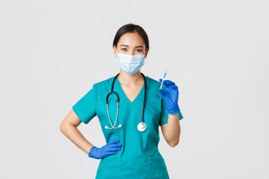 Covid-19, koronavirüs hastalığı, sağlık çalışanları konsepti. Önlüklü ve tıbbi maskeli gülümseyen, kendine güvenen Asyalı kadın hemşire, elinde aşı şırıngası, aşı aşısı, beyaz arka plan