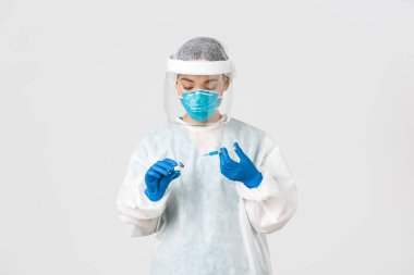 Covid-19, koronavirüs hastalığı, sağlık çalışanları konsepti. Ciddi görünümlü kadın hekim, kişisel koruma ekipmanlarında Asyalı doktor aşıyla birlikte ampul içine şırınga yerleştirin.