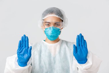 Covid-19, koronavirüs hastalığı, sağlık çalışanları konsepti. Ciddi görünümlü endişeli Asyalı kadın doktorlara yakın çekim. Kişisel koruma ekipmanlarında dur hareketi göster, tehlikeli bölgeye girmeni yasakla.