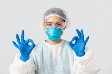 Covid-19, koronavirüs hastalığı, sağlık çalışanları konsepti. Kişisel koruma ekipmanlarıyla memnun olmuş, ciddi görünümlü Asyalı bir kadının yakın plan çekimi tamam bir jest yap, onayla ya da garanti et