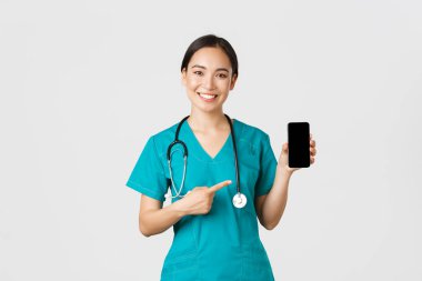 Covid-19, sağlık çalışanları ve online tıp konsepti. Gülümseyen Asyalı kadın doktor, ameliyat önlüklü doktor, hareket halindeki ekranı işaret ediyor, check-up 'ı indirmeyi, danışmanlık uygulamasını öneriyor