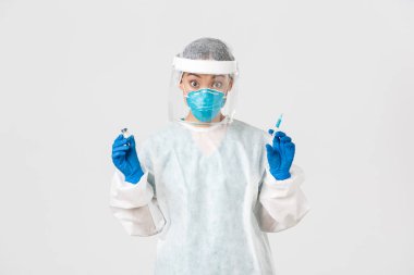 Covid-19, koronavirüs hastalığı, sağlık çalışanları konsepti. Şaşırmış ve heyecanlı Asyalı doktor, kişisel koruyucu ekipman enfeksiyonisti, elinde şırınga ve aşı ampulü var.