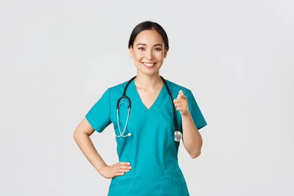 Covid-19, profissionais de saúde, conceito pandémico. Confiante sorrindo asiático profissional médico, enfermeira em esfrega apontando o dedo para a câmera, convidando para check-up ou vacinação, dar recomendação — Fotografia de Stock