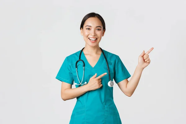 Covid-19, profissionais de saúde, conceito pandémico. Sorrindo alegre asiático feminino médico, terapeuta em esfrega apontando dedos canto superior direito, convidando para check-up, recomendo vacinar — Fotografia de Stock