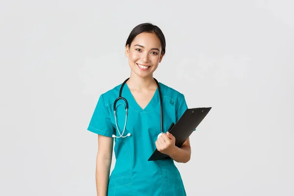 Healthcare workers, preventing virus, quarantine campaign concept. Smiling asian female nurse, doctor with clipboard wearing scrubs, provide checkup, examine patient in clinic, white background — Stock Photo, Image