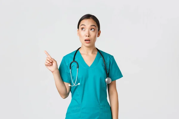 Covid-19, pracownicy służby zdrowia, koncepcja pandemii. Zaskoczona i zdumiona kobieta azjatycki lekarz patrząc i wskazując lewy górny róg. Zdumiewający koreański lekarz gapi się na baner z reklamą — Zdjęcie stockowe