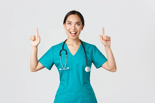 Covid-19, egészségügyi dolgozók, világjárvány. Szórakoztató vidám ázsiai női nővér, orvos vagy sebész műtősruhák ujjal mutogató és felnézett elégedett arc, álló fehér háttér — Stock Fotó