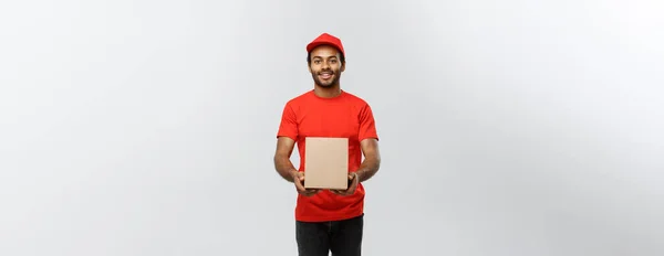 Αντίληψη παράδοσης - Πορτρέτο του Happy African American delivery man σε κόκκινο πανί κρατώντας ένα πακέτο κουτί. Απομονωμένος στο Grey studio Background. Αντιγραφή χώρου. — Φωτογραφία Αρχείου
