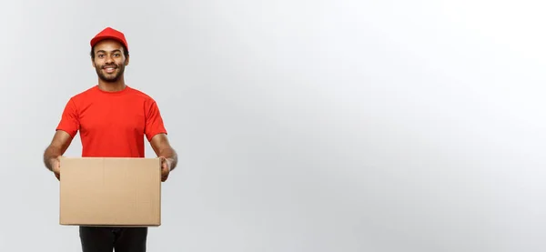 Conceito de entrega - Retrato de homem de entrega afro-americano feliz em pano vermelho segurando um pacote de caixa. Isolado no estúdio Grey Background. Espaço de cópia . — Fotografia de Stock