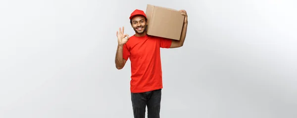 Doručení koncepce - portrét Happy African American doručovatel držení box balení a ok známek. Izolované na šedé studio pozadí. Kopírovat prostor. — Stock fotografie