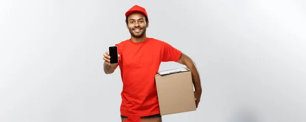 Leverans koncept - porträtt av stilig African American leverans mannen eller courier med box visar mobiltelefon på dig att kontrollera ordningen. Isolerad på grå studio bakgrund. Kopiera utrymme. — Stockfoto