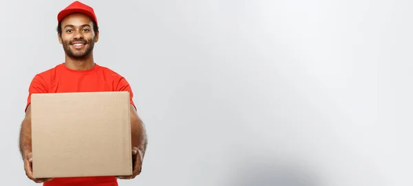 Delivery Concept - portré a boldog afro-amerikai kézbesítő férfi piros ruhával kezében egy doboz csomagot. Elszigetelve a szürke stúdió hátterében. Fénymásolási tér. — Stock Fotó