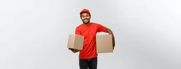 Lieferkonzept - Porträt eines glücklichen afroamerikanischen Zustellers in rotem Tuch, der ein Karton-Paket hält. Isoliert auf grauem Studiohintergrund. Kopierraum. — Stockfoto