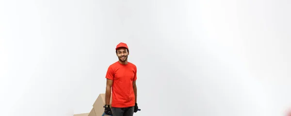 Concepto de entrega - Retrato del guapo repartidor afroamericano o mensajero empujando un camión de mano con una pila de cajas. Aislado en Grey studio Background. Copiar espacio . —  Fotos de Stock