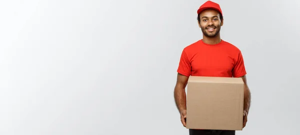 Conceito de entrega - Retrato de homem de entrega afro-americano feliz em pano vermelho segurando um pacote de caixa. Isolado no estúdio Grey Background. Espaço de cópia . — Fotografia de Stock