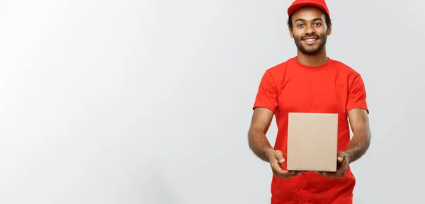 Lieferkonzept - Porträt eines glücklichen afroamerikanischen Zustellers in rotem Tuch, der ein Karton-Paket hält. Isoliert auf grauem Studiohintergrund. Kopierraum. — Stockfoto