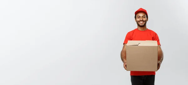 Concept de livraison - Portrait du livreur afro-américain heureux en tissu rouge tenant un paquet de boîte. Isolé sur fond de studio Grey. Espace de copie. — Photo