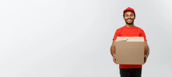 Concepto de entrega - Retrato del hombre de entrega afroamericano feliz en tela roja sosteniendo un paquete de caja. Aislado en Grey studio Background. Copiar espacio . —  Fotos de Stock