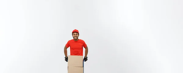 Concepto de entrega - Retrato del guapo repartidor afroamericano o mensajero empujando un camión de mano con una pila de cajas. Aislado en Grey studio Background. Copiar espacio . — Foto de Stock