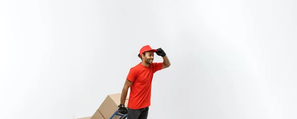 Concepto de entrega - Retrato del guapo repartidor afroamericano o mensajero empujando un camión de mano con una pila de cajas. Aislado en Grey studio Background. Copiar espacio . —  Fotos de Stock