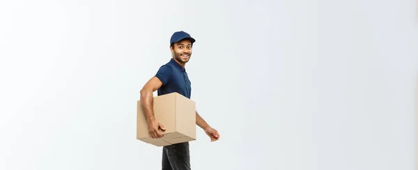 Lieferkonzept - Porträt eines glücklichen afrikanisch-amerikanischen Zustellers in blauem Tuch, der zu Fuß ein Karton-Paket an den Kunden schickt. isoliert auf grauem Studiohintergrund. Kopierraum. — Stockfoto