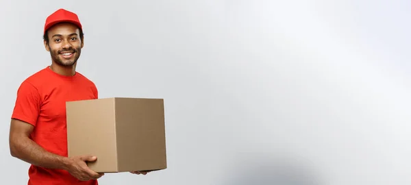 Lieferkonzept - Porträt eines glücklichen afroamerikanischen Zustellers in rotem Tuch, der ein Karton-Paket hält. Isoliert auf grauem Studiohintergrund. Kopierraum. — Stockfoto