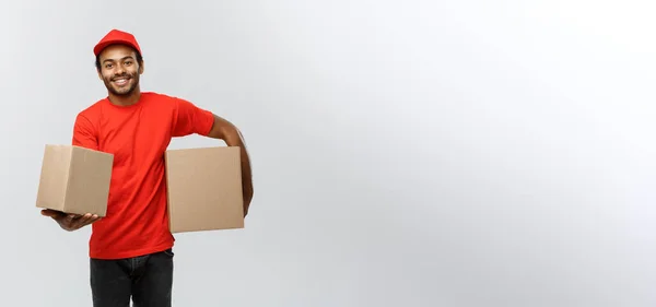 Conceito de entrega - Retrato de homem de entrega afro-americano feliz em pano vermelho segurando um pacote de caixa. Isolado no estúdio Grey Background. Espaço de cópia . — Fotografia de Stock