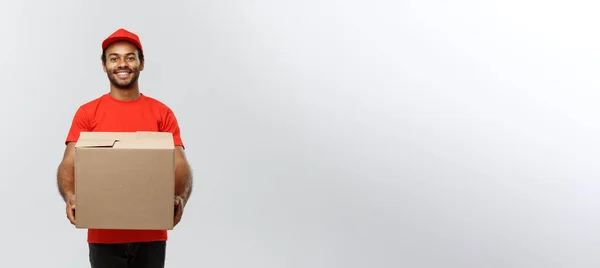 Concepto de entrega - Retrato del hombre de entrega afroamericano feliz en tela roja sosteniendo un paquete de caja. Aislado en Grey studio Background. Copiar espacio . —  Fotos de Stock