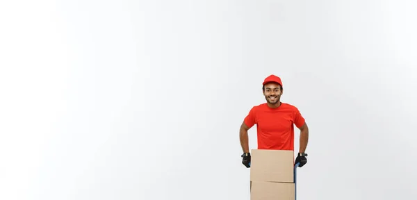 Concepto de entrega - Retrato del guapo repartidor afroamericano o mensajero empujando un camión de mano con una pila de cajas. Aislado en Grey studio Background. Copiar espacio . — Foto de Stock