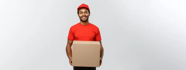 Concepto de entrega - Retrato del hombre de entrega afroamericano feliz en tela roja sosteniendo un paquete de caja. Aislado en Grey studio Background. Copiar espacio . — Foto de Stock