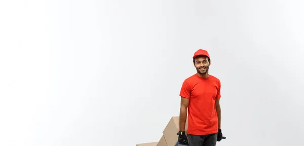 Concepto de entrega - Retrato del guapo repartidor afroamericano o mensajero empujando un camión de mano con una pila de cajas. Aislado en Grey studio Background. Copiar espacio . —  Fotos de Stock